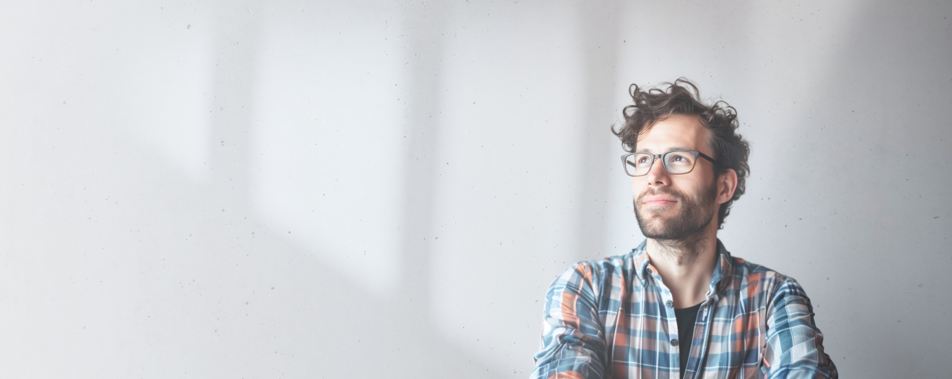 A man with glasses and curly hair is looking thoughtfully to the side. He is wearing a plaid shirt and stands against a plain light gray wall with soft shadows.