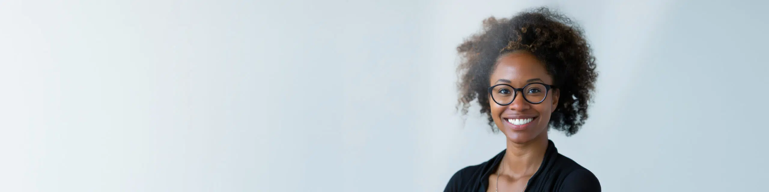 A person with curly hair and glasses smiles warmly at the camera, standing against a light grey background. They are wearing a black top and a small earring is visible.