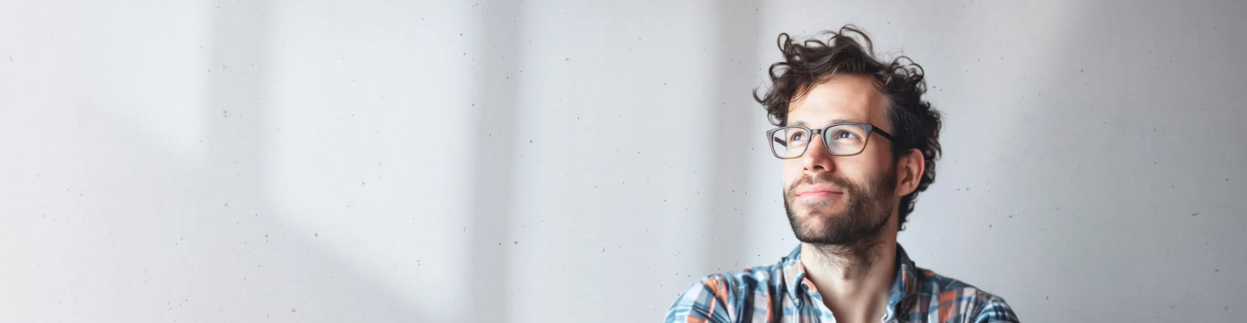 A man with curly hair, glasses, and a beard wearing a plaid shirt looks thoughtful while gazing upward, standing against a light gray background.